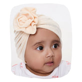 Beige Baby Turban Cap with shiny finish and floral knot, worn by infant girl. Ultra-soft, stretchable design for 0 to 2 years.